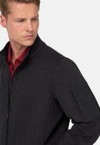 Črna zip-up jakna z visokim ovratnikom, gladko teksturo in nalepko na levem rokavu z kontrastnimi tkaninami.