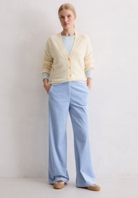 Cremefarbener Cardigan mit V-Ausschnitt und Knopfleiste, darüber getragen ein hellblau gestreiftes Langarmshirt und weite hellblaue Hosen.