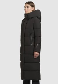 Frau in einem langen schwarzen gepolsterten Wintermantel mit hohem Kragen und Kapuze, seitlichen Reißverschluss-Taschen, steht vor einem einfarbigen Hintergrund.