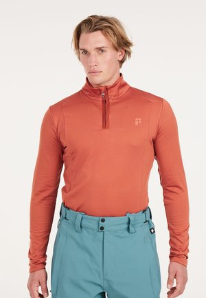 Langarm, kwart rits bovenstuk in roest-oranje stof, met een staande kraag en een gladde textuur, gecombineerd met lichtblauwe broek.