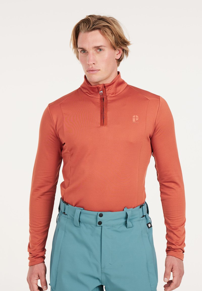 Langarm, kwart rits bovenstuk in roest-oranje stof, met een staande kraag en een gladde textuur, gecombineerd met lichtblauwe broek.