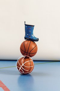 Sininen kuminen sadetakki, jossa on leikillisiä grafiikoita, istuu kahden pinotun oranssin koripallon päällä sinisellä liikuntasalilattialla, jossa on valkoinen koripallon ääriviiva.