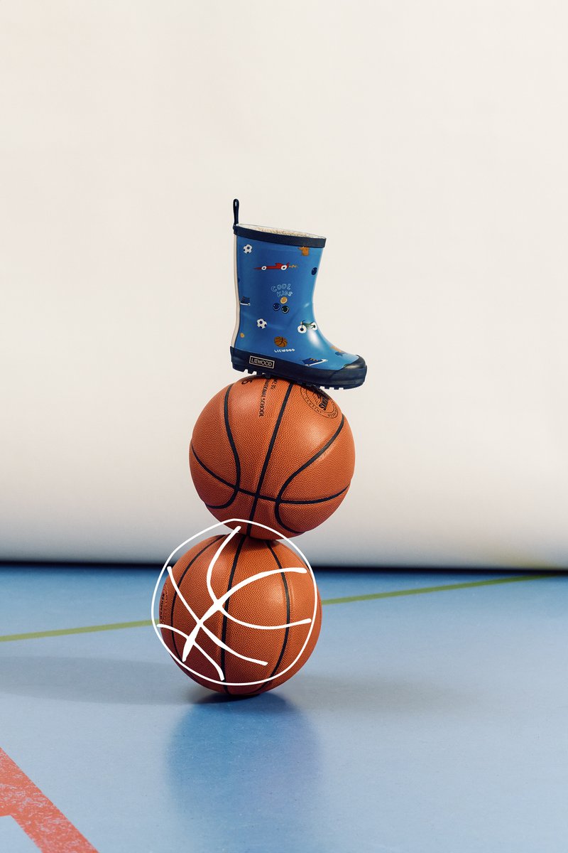 Sininen kuminen sadetakki, jossa on leikillisiä grafiikoita, istuu kahden pinotun oranssin koripallon päällä sinisellä liikuntasalilattialla, jossa on valkoinen koripallon ääriviiva.