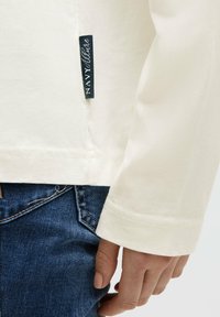 Cremefarbenes Langarm-T-Shirt mit einem marineblauen Etikett, kombiniert mit dunkelblauen Jeans. Beide Stoffe wirken weich und lässig im Design.