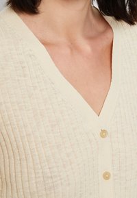 Cárdigan crema con diseño de escote en V, textura de canalé y dos botones beige en la parte delantera. El material parece ligero y ligeramente translúcido.