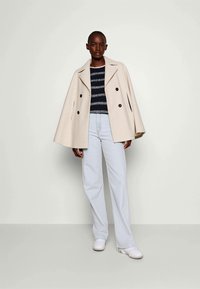 Donna che indossa un cappotto beige a doppio petto sopra un maglione a righe bianche e nere, con jeans a gamba larga azzurri e sneakers bianche.