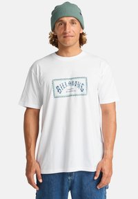 Hvid bomulds T-shirt med blåt "Billabong" logo med dekorativ kant; parret med blå denim shorts og en grå strikket beanie.