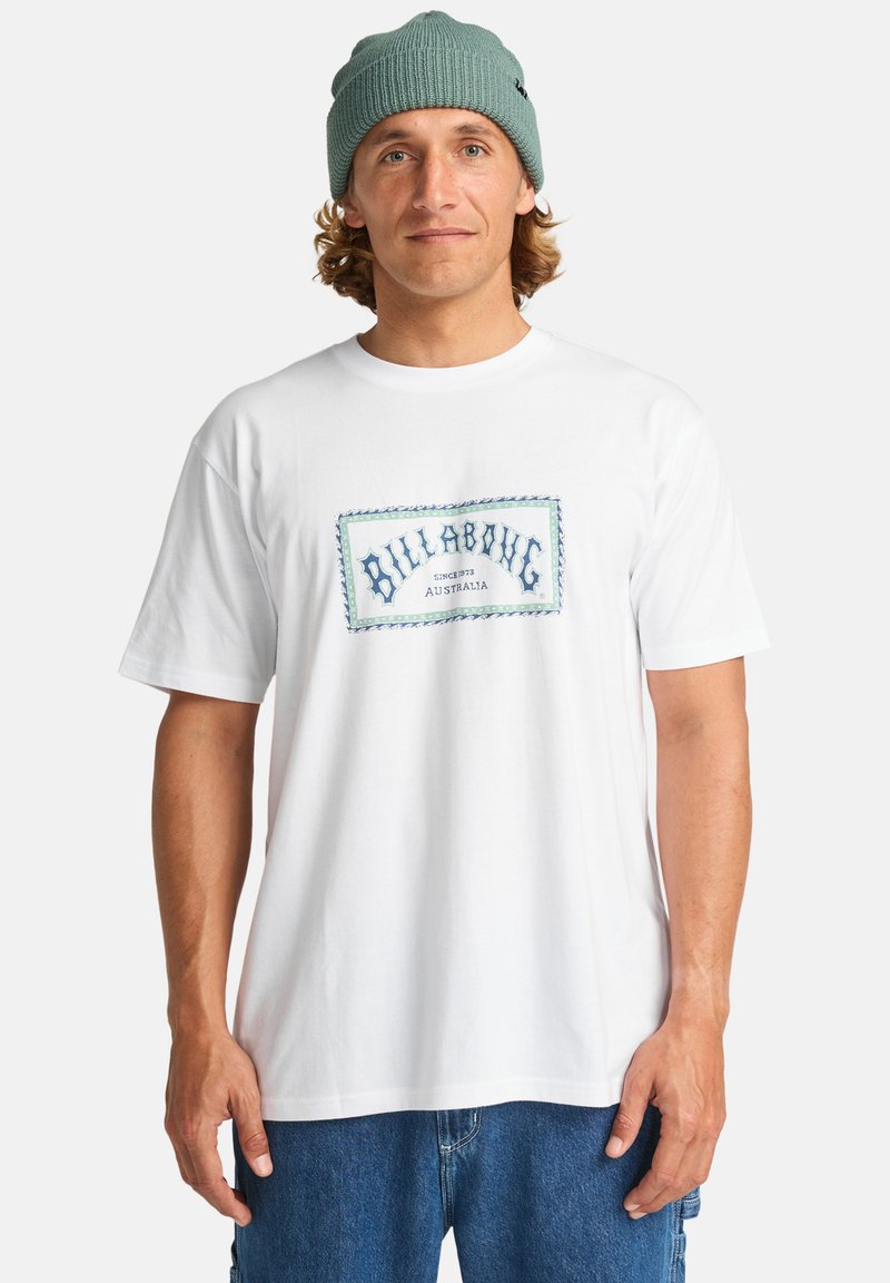 T-shirt bianca in cotone con logo blu "Billabong" e bordo decorativo; abbinata a pantaloni corti di denim blu e un berretto in maglia grigia.