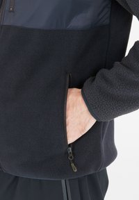 Fleecejakke i mørk navy, med en glat nylon øverste sektion, lynlåslommer og tekstureret stof. Nærbillede af hånd i lommen.