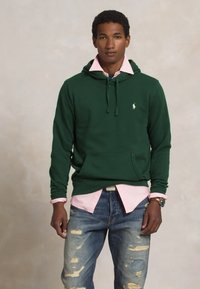Groene hoodie met een voorzak en trekkoord, gelaagd over een roze overhemd met kraag, gecombineerd met versleten blauwe jeans.