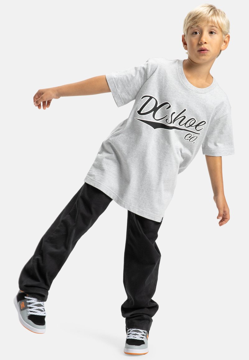 Šedé tričko s krátkým rukávem a černým logem "DC shoe co.", v kombinaci s černými volnými kalhotami a černobílými teniskami s oranžovými akcenty.