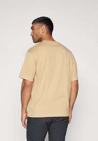 Tricou din bumbac bej cu mâneci scurte, gât rotund și un logo mic imprimat în partea superioară a spatelui. Textură netedă, croi casual.