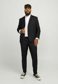 Schwarzer Anzug mit einem taillierten Blazer und passender Hose, kombiniert mit einem weißen Hemd und schwarzen Sneakers, auf einem neutralen Hintergrund präsentiert.
