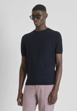 Man draagt een zwart kortärmelig truitje en lichtroze broek, staat met ontspannen armen en draagt een zwarte zonnebril tegen een grijze achtergrond.