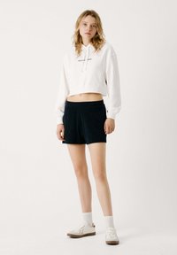 Bijela skraćena hoodie s pletenicom i otisnutim tekstom, uparen s crnim sportskim šortsevima. Model nosi bijele tenisice i čarape do gležnja.