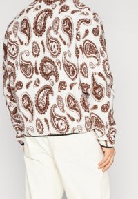 Osoba v bílém fleecovém svetru s hnědými paisley vzory a krémových kalhotách, zobrazená zezadu s jednou rukou u boku.