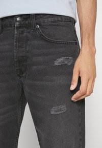 Vaqueros de mezclilla negra con corte slim, que cuentan con desgastes y rotos en los muslos, diseño de cinco bolsillos y cierre clásico de botón y cremallera.