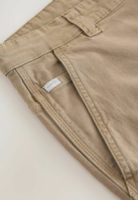 Pantalons en coton beige avec une texture lisse et une coupe droite. Présentent une boucle de ceinture et une étiquette cousue sur la taille.