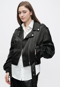 Čierna saténová krátka bunda so zipsom a zapínaním na cvoky, nafúknutými rukávmi, bočnými vreckami a nastaviteľnými detailmi na popruhoch. Kombinovaná s bielou košeľou.