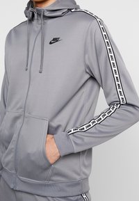 Šedá mikina s kapucí na zip, s černým logem Nike na hrudi a bílým stuhou s logem po stranách rukávů. Měkčí materiál, volný střih.