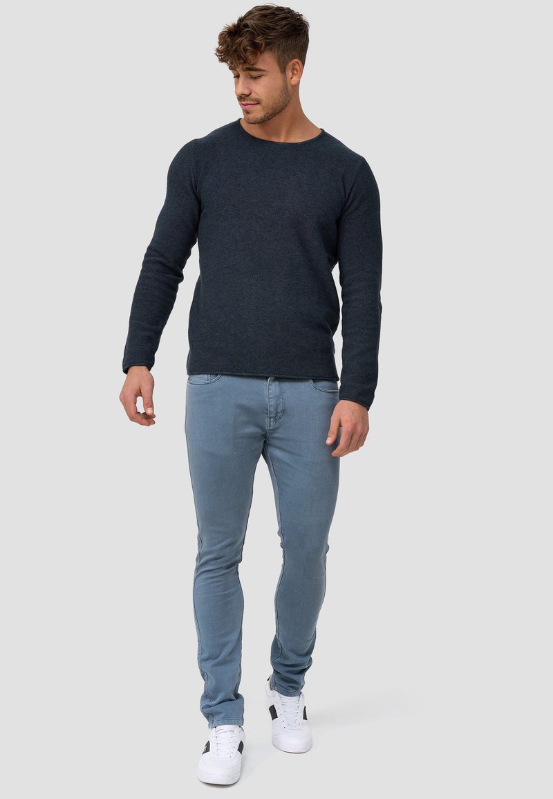 Jeune homme aux cheveux bouclés portant une chemise à manches longues foncée, un jean slim bleu clair et des baskets blanches, debout sur un fond uni.