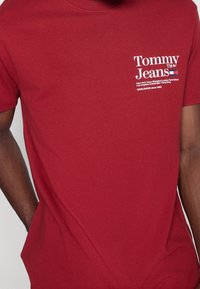 T-shirt en coton rouge à manches courtes, avec un design de logo blanc sur la poitrine incluant du texte et des noms de villes internationales.
