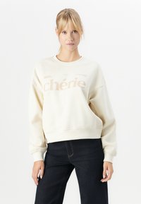 Joven mujer con cabello rubio en una cola de caballo lleva un sudadera color crema con el texto "chérie" y jeans azul oscuro, de pie contra un fondo blanco.
