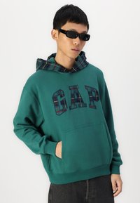 Pulover verde cu glugă căptușită cu carouri, având un logo mare "GAP" în culori contrastante. Material moale, buzunar frontal și croială lejeră.