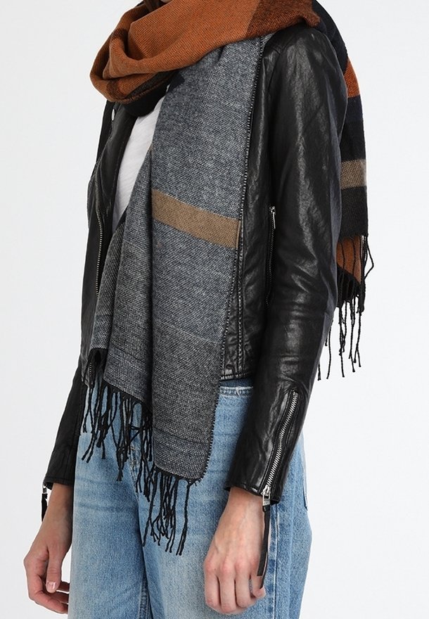 Personne portant une veste en cuir noire, un jean bleu et une grande écharpe bicolore gris et marron avec des extrémités à franges.