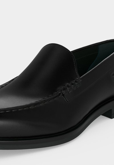 Μαύρος δερμάτινος loafer τύπου slip-on με ραφές στιλ μοκασίν και χαμηλό στιβαρό τακούνι, εμφανίζεται σε λευκό φόντο.