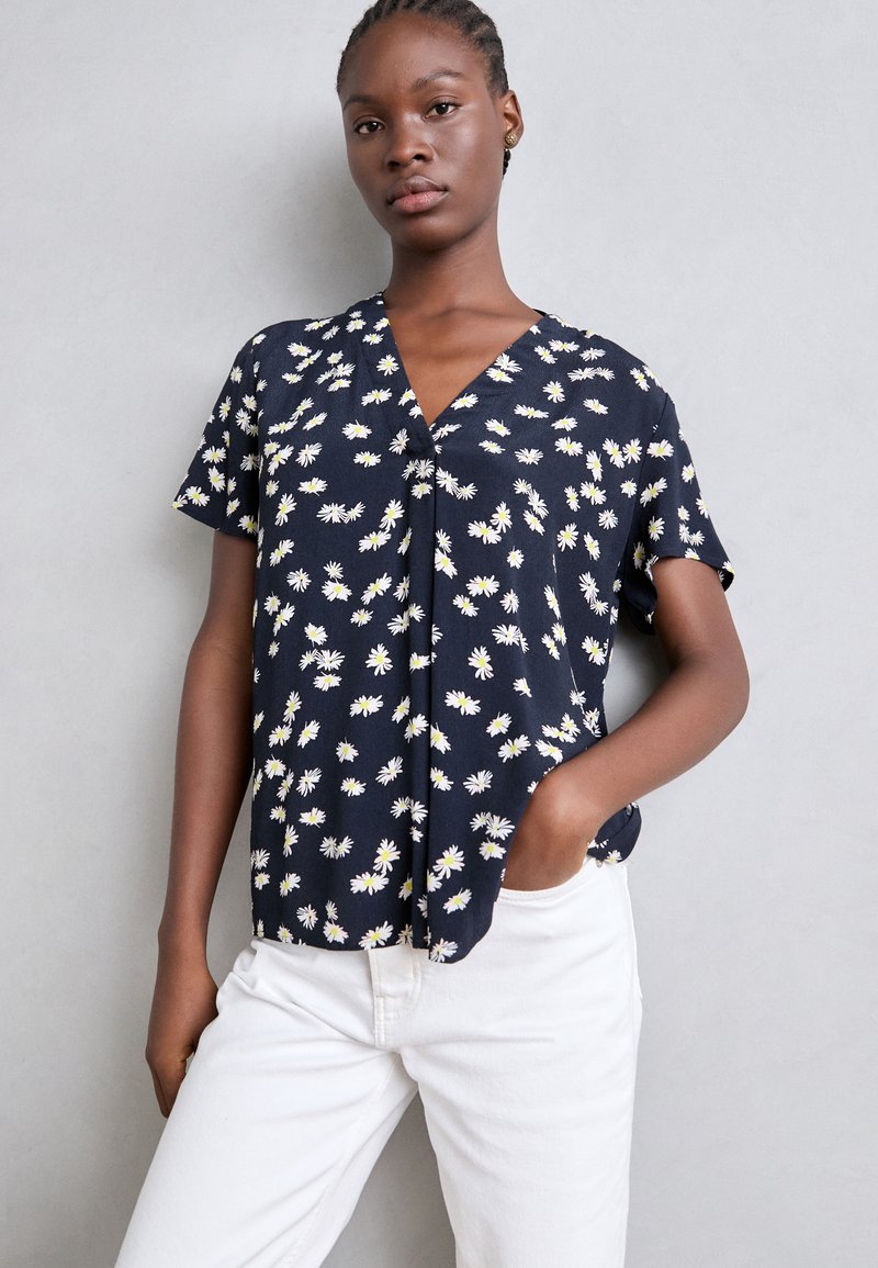 Blusa a maniche corte blu navy con motivo floreale bianco e giallo, design a scollo a V e vestibilità rilassata. Il tessuto sembra leggero e strutturato.
