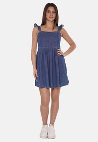 Abito in denim con spalline a volant regolabili, scollatura quadrata e gonna svasata. Colore: blu scuro. Indossato con sneaker bianche.