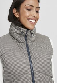 Kvadrični pufasti prsluk u kariranom uzorku bež i crne boje. Ima visoki ovratnik, mornarski zatvarač i podesivu šnalu.