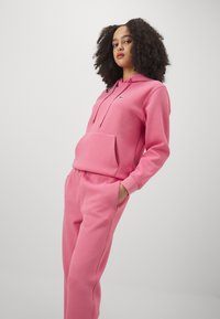 Felpa rosa con pantaloni coordinati realizzati in tessuto morbido, con tasca frontale e cordini regolabili. Dettaglio del logo ricamato.