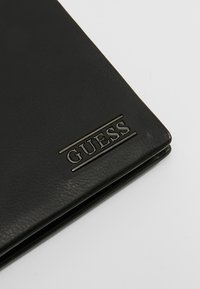 Portafoglio in pelle nera con logo "GUESS" in rilievo. Texture liscia con design elegante, bordi piegati e stile minimalista.