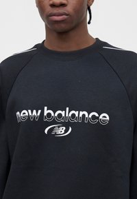 Άτομο που φοράει μαύρο φούτερ New Balance με λευκό λογότυπο στο στήθος, περικομμένο στο κάτω μέρος του προσώπου και τους ώμους.