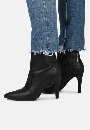 Schwarze Leder-Ankle-Boots mit spitzer Zehenform und Stiletto-Absatz, die eine glatte Textur und einen seitlichen Reißverschluss haben. Die Jeans haben einen fransigen Saum.