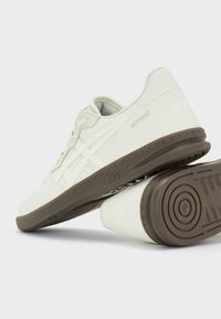 Baskets blanches Asics SkyHand avec semelles marron, logo latéral et motifs de grip texturés sous la semelle, présentées inclinées sur fond blanc.