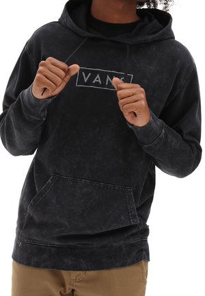 Personne portant un hoodie Vans noir délavé, tenant les cordons du hoodie avec les deux mains, portant un pantalon beige.