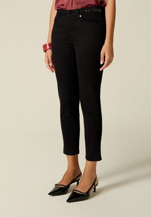 Nainen, jolla on mustat cropped-tasaiset slim-fit-housut, mustat kärkikärkiset slingback-korkokengät ja punainen toppi, jossa on ranteessa sopiva rannekoru.