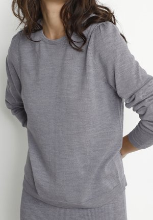 Kvinde med bølget brunt hår iført en lysegrå strikket trøje med lange ærmer og matchende ribstrikket nederdel, stående med hænderne bag ryggen.