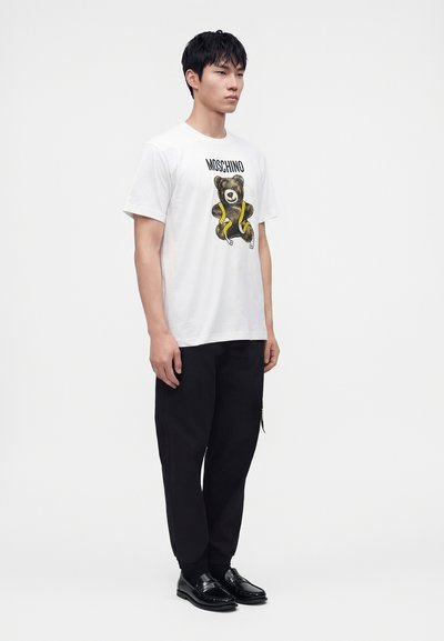 Νέος άνδρας στέκεται φορώντας ένα λευκό t-shirt Moschino με σχέδιο αρκούδας και ταινίας, μαύρο παντελόνι και μαύρα loafers σε απλό φόντο.