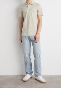 Polo beige avec un petit logo, associé à un jean bleu clair avec un détail usé, et des baskets blanches avec des semelles marron.