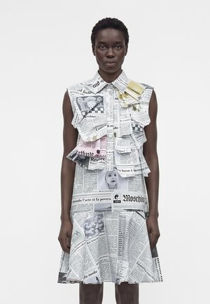 Naine kannab varrukateta kraega kleiti, millel on kihilised ajalehepildid ja pildid ühetaustalisel heledal taustal.