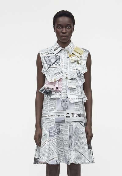 Woman wearing a sleeveless, collared dress printed with layered newspaper pages and images against a plain light background.