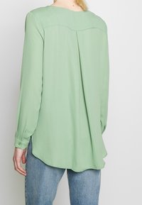 Camicia a maniche lunghe in tessuto verde chiaro, caratterizzata da uno scollo arrotondato, dettaglio piega posteriore e vestibilità ampia con orlo curvo. Abbinata a jeans blu.