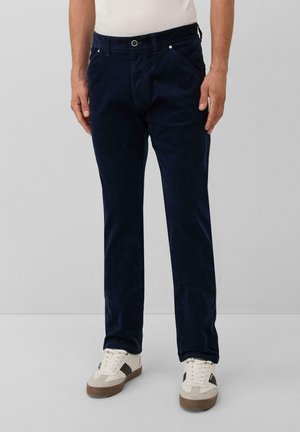Mann in dunkelblauen Cordhosen und weißen Sneakers mit schwarzen und braunen Details, der vor einem schlichten grauen Hintergrund steht.