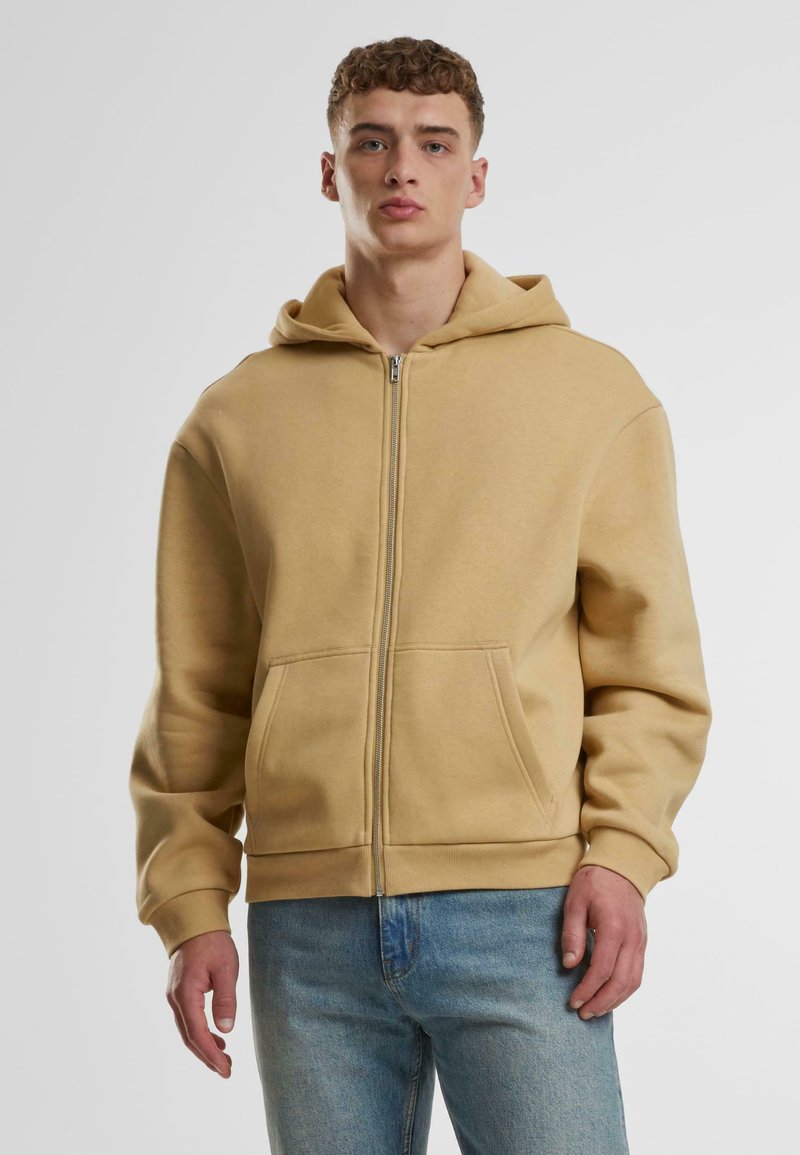 Beige zip-up hoodie med en romslig lomme foran, myk tekstur og justerbar hette. Brukt over blå jeans, med ribbestrikkede mansjetter og kant.