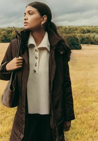 Pruun isoleeritud jakk kõrge kraega, kreemjas kudumiskampsun nelja nööbiga ja hall käekott. Loomulik välistingimustes.