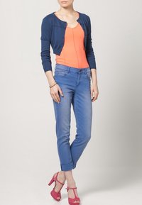 Tajtat marinblå cardigan, klarorange topp, ljusblå jeans med uppvikt fåll och rosa klackar med remmar. Slät tyg och avslappnad design.